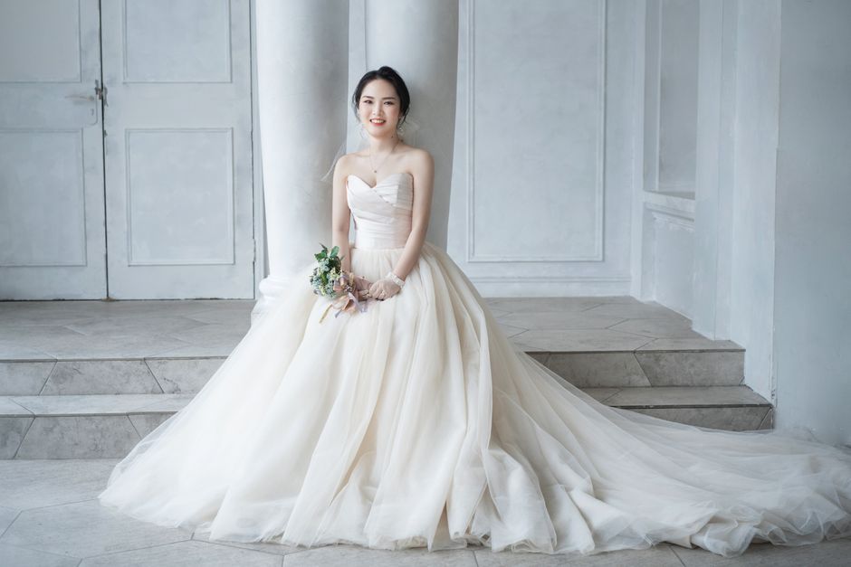 美しい花嫁が流れるウェディングドレスを着て、小さなブーケを持って室内に座っています。