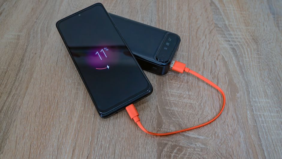 木製のテーブルの上で、オレンジ色のケーブルを使ってモバイルバッテリーから充電されているスマートフォン。