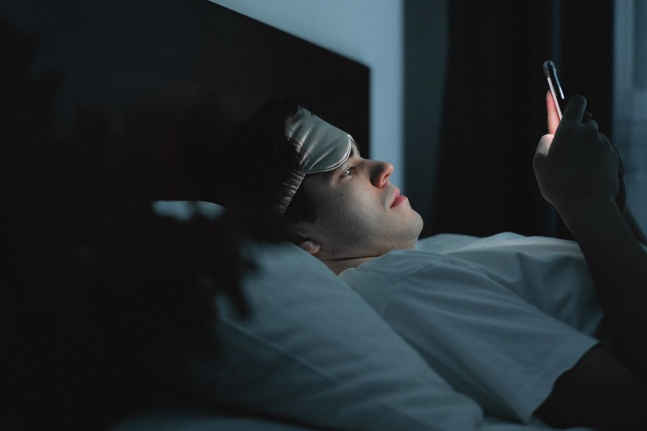 睡眠マスクを着用した男性が、夜遅くにベッドでスマートフォンを使用しています。